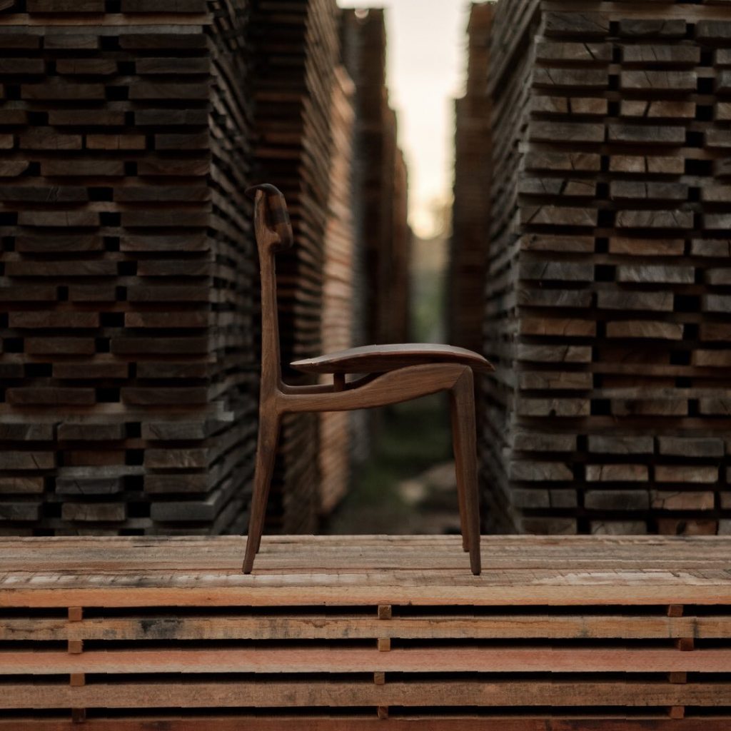 Designer chair made from sustainable Brazilian wood, displayed in a rustic setting with stacked hardwood planks in the background.